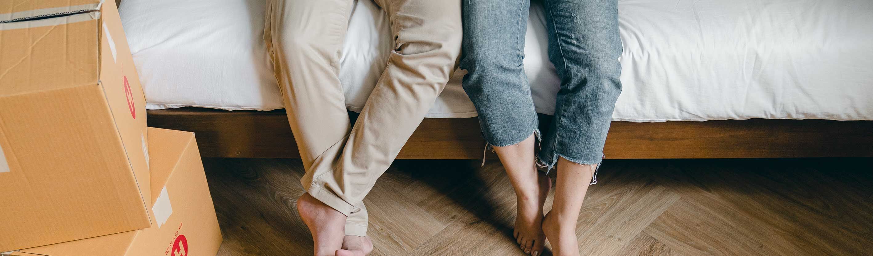 Bildausschnitt zeigt Paar auf Bett sitzend mit Füßen herabbaumelnd zum Holzboden.